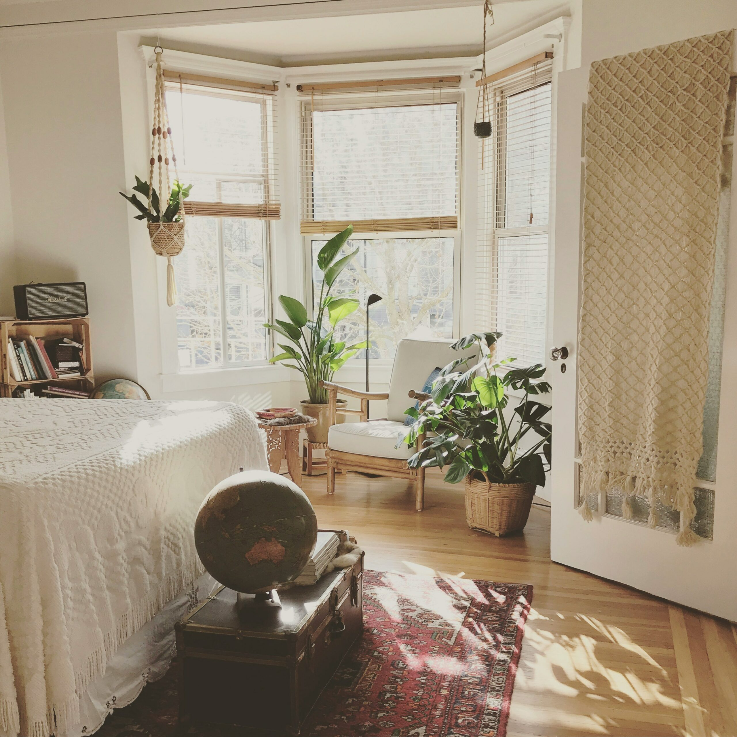 Bedroom with plants, a rug, a wooden chair, and wooden blinds lifted up allowing sun to enter